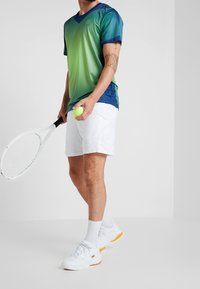 Tričko s krátkým rukávem v zeleno-modrém gradientu, bílé šortky, držící tenisový míček a raketu, v bílých teniskách a ponožkách.