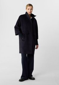 Marineblauer Wollmischungs-Mantel mit hohem Kragen, tiefen Schultern, zwei vorderen aufgenähten Taschen und einer lässigen Passform, kombiniert mit dunklen weiten Hosen.
