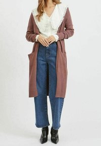 Lång, brun kofta med fickor, bärs över en vit blus med spetskrage, ihopparad med högmidjade blå jeans och svarta stövlar.