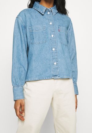 Osoba ubrana w jasnoniebieską, dżinsową koszulę na guziki z kieszeniami na piersi oraz białe spodnie, stojąca na jednolitym tle.