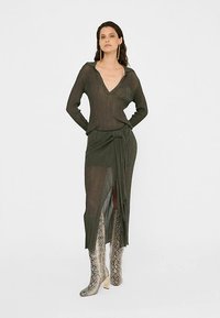 Olivgrünes Strickkleid mit langen Ärmeln, tiefem V-Ausschnitt und seitlichem Schlitz. Strukturierter Stoff, tailliert, kombiniert mit metallisch schimmernden Stiefeln im Schlangenledermuster.