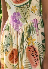 Kukallinen mekko, jossa on suuria vesimeloneja, viinirypäleitä ja lehtiä vihreän, vaaleanpunaisen, violetin ja keltaisen sävyissä, ja rypytetty vyötärö.