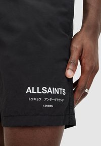 Black shorts made of lightweight fabric. Features white printed text "ALLSAINTS," symbols, and "LONDON" at the hem.
