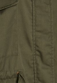 Tkanina oliwkowa o fakturowanym wykończeniu, z boczną kieszenią i sznurkiem zabezpieczonym mosiężnym przelotem.