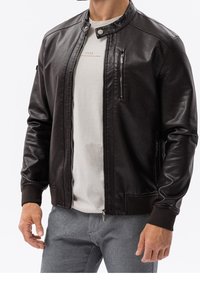 Muž oblečený v tmavě hnědé kožené bundě přes světle béžové tričko a šedé kalhoty, stojící s uvolněnýma rukama podél těla.