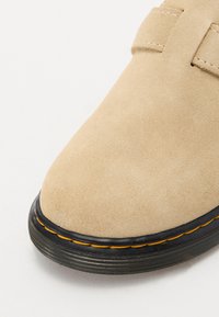 Beige semskede sko med rund tå, som har en sort teksturert såle og en oransje dekorativ søm langs kanten.