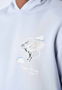 Hoodie bleu clair avec un graphique de cheval ailé en blanc et gris clair, accompagné de texte et de motifs de nuages imprimés en dessous. Tissu au toucher doux.