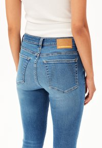 Une personne portant un jean en denim bleu clair avec des poches arrière et une étiquette marron "ARMEDANGELS", associé à un haut sans manches blanc.