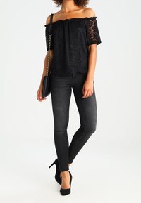 Schwarzes Spitzen-Oberteil ohne Ärmel mit kurzen Ärmeln, kombiniert mit eng anliegenden schwarzen Jeans und schwarzen High Heels, dazu eine schwarze Umhängetasche.