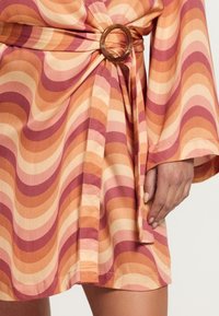 Primer plano de una mano descansando junto a un vestido mini con un patrón ondulado en naranja, rosa y crema, y una hebilla circular marrón.