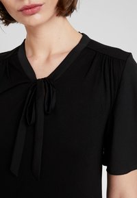 Svart blus med V-ringning, korta ärmar och en knytning vid halsringningen, tillverkad av mjukt tyg med slät textur.