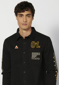 Camisa negra de botones con cuello, que presenta gráficos amarillos, un bolsillo en el pecho con texto blanco y un parche en forma de triángulo en el hombro.