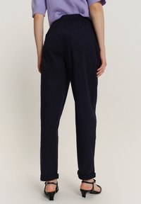 Persoon in marineblauwe hoge taillebroek met elastische tailleband, opgerolde manchetten, gecombineerd met zwarte strappy laaghaks, gezien vanachter.