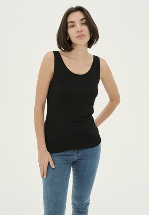 Žena s krátkymi tmavými vlasmi, oblečená v čiernom tričku bez rukávov a modrých džínsoch, stojaca s jednou rukou na bok proti bielemu pozadiu.