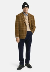 Mann trägt eine schwarze Mütze, ein braunes kariertes Blazer, einen beigen Strickpullover über einem blauen Hemd, eine marineblaue Cordhose und schwarze Schnürschuhe und steht mit einer Hand in der Tasche.