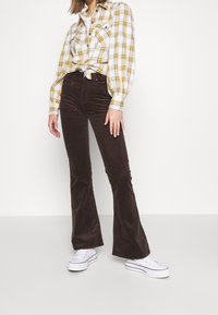 Femme portant une chemise à carreaux blanche et jaune nouée à la taille, un pantalon en velours côtelé marron foncé évasé, et des baskets blanches à plateforme, faisant face à un fond blanc.