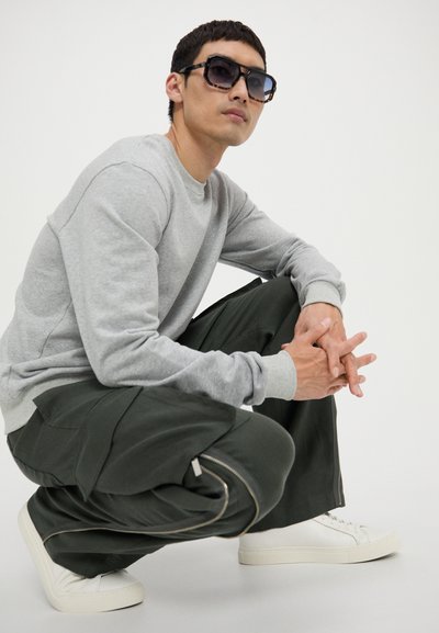 Gray sweatshirt, dark green cargo pants with side zippers, white sneakers, and bold square sunglasses. Model crouching, hands clasped.