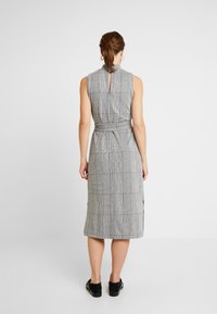 Vestido sin mangas de longitud hasta la rodilla en un patrón de cuadros en blanco y negro, con un cuello alto, detalle de ojal en la parte posterior y cinturón en la cintura.
