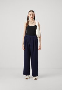 Black tank top, navy wide-leg trousers with pleats, and white shoulder bag. Model wears white sneakers, standing on a light background.