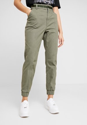 Pantalons tapered en mélange de coton de couleur khaki, dotés d'une taille haute, de poignet élastiques et de poches latérales, associés à des baskets blanches.
