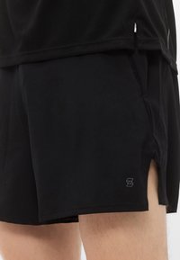 Μαύρο αθλητικό σορτς από ελαφρύ ύφασμα, με πλάγιες σκισμές, ελαστική μέση και μικρό λογότυπο στο κάτω μέρος.