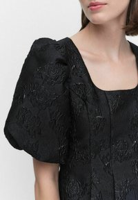 Svart klänning med stora puffärmar, prydd med texturerad blomstersbroderi. Fyrkantig halsringning och slät tyg med glans.