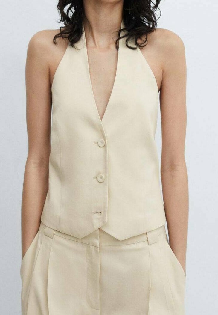 Veste beige sans manches avec un décolleté en V profond, dotée de deux boutons et d'une coupe ajustée, fabriquée en tissu lisse. Stylisée avec un pantalon assorti taille haute.