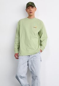 Šviesiai žalia ilgomis rankovėmis marškinėliai su Carhartt logotipu, derinami su šviesiai pilkomis džinsomis. Modelis dėvi suderinamą žalią kepurę. Paprastas dizainas.