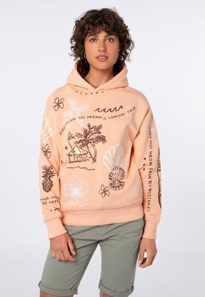 Woman wearing a peach hoodie with tropical drawings and motivational text, paired with rolled-up olive green shorts, standing against white background.