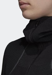 Haut noir à fermeture éclair avec un tissu texturé côtelé. Il présente un col montant et des accents réfléchissants subtils le long de la fermeture éclair et des coutures.