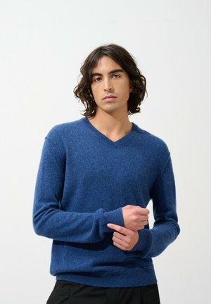 Niebieski sweter w serek wykonany z miękkiego dzianiny, z prążkowanymi mankietami i dołem; model nosi go w pomieszczeniu na tle gładkiej ściany.