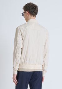 Beige lichtgewicht jas met elastische manchetten en tailleband, met een opstaande kraag en een gladde textuur, gezien vanuit de achterkant.