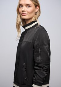 Veste bomber noire avec fermeture éclair, col et poignets côtelés. Dispose de poches latérales et d'une poche zippée sur la manche. Texture lisse.