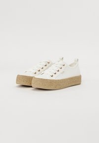 Hvite lerretssko med en teksturert, vevd jutesåle, med gulløyeblikk og flate lisser. Slip-on-stil med en løkke på hælen.