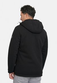 Chaqueta negra con capucha hecha de tela texturizada, con cordones ajustables y un detalle de logo en la manga, combinada con pantalones de color claro.