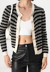 Kobieta ubrana w czarno-białą, prążkowaną kardigan z dzianiny, nosząca białą bluzkę na crop top oraz czarne skórzane spodnie i białą torebkę na ramię.