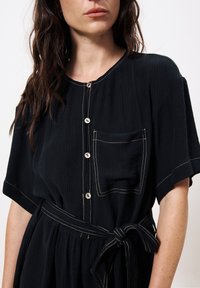 Das schwarze knöpfbare Kleid verfügt über kurze Ärmel, eine Tasche mit kontrastierender Naht und eine gebundene Taille für eine verstellbare Passform. Strukturierter Stoff.