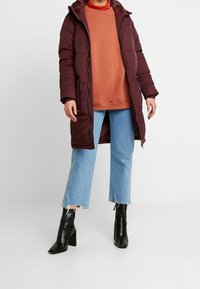 Cappotto puffer bordeaux con cappuccio, caratterizzato da cuciture verticali, chiusura frontale con zip, indossato su una felpa arancione e jeans cropped blu, abbinato a stivali neri alla caviglia.