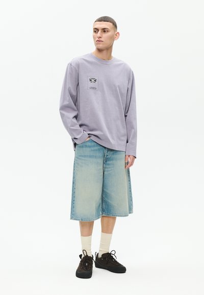 Jeune homme debout, portant une chemise à manches longues lavande avec un petit motif graphique, un short en denim large bleu clair, des chaussettes beiges et des baskets noires.