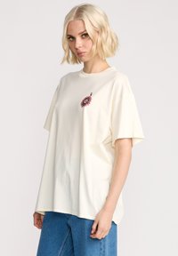 T-shirt en coton surdimensionné de couleur crème avec un graphique de fleur rose sur la poitrine. Manches courtes et coupe décontractée.