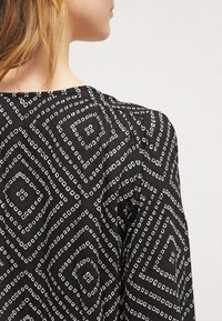 Černá blůza s bílým geometrickým diamantovým vzorem. Materiál se zdá být lehký s splývavou texturou a zaobleným výstřihem.