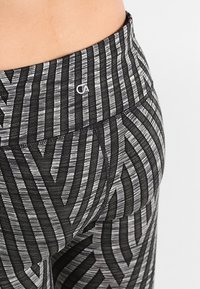 Zbliżenie na osobę noszącą legginsy z wysokim stanem w czarno-szare geometryczne paski oraz małym białym logo na pasku.