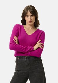 Sweter w serek w intensywnym różowym kolorze, wykonany z miękkiej dzianiny. Długie rękawy z dopasowanymi mankietami oraz luźny fason, zestawiony z ciemnymi spodniami o wysokim stanie.