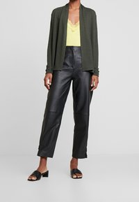 Olivgrüner Cardigan über einem gelben Spitzenoberteil, kombiniert mit schwarzen, hoch taillierten Lederhosen und schwarzen Sandalen mit Absatz.