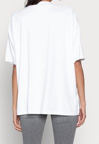 Bílá oversized tričko s krátkými rukávy, kulatým výstřihem a bočními rozparky. Hladká tkanina s volným střihem. Šedé slim-fit kalhoty viditelné.