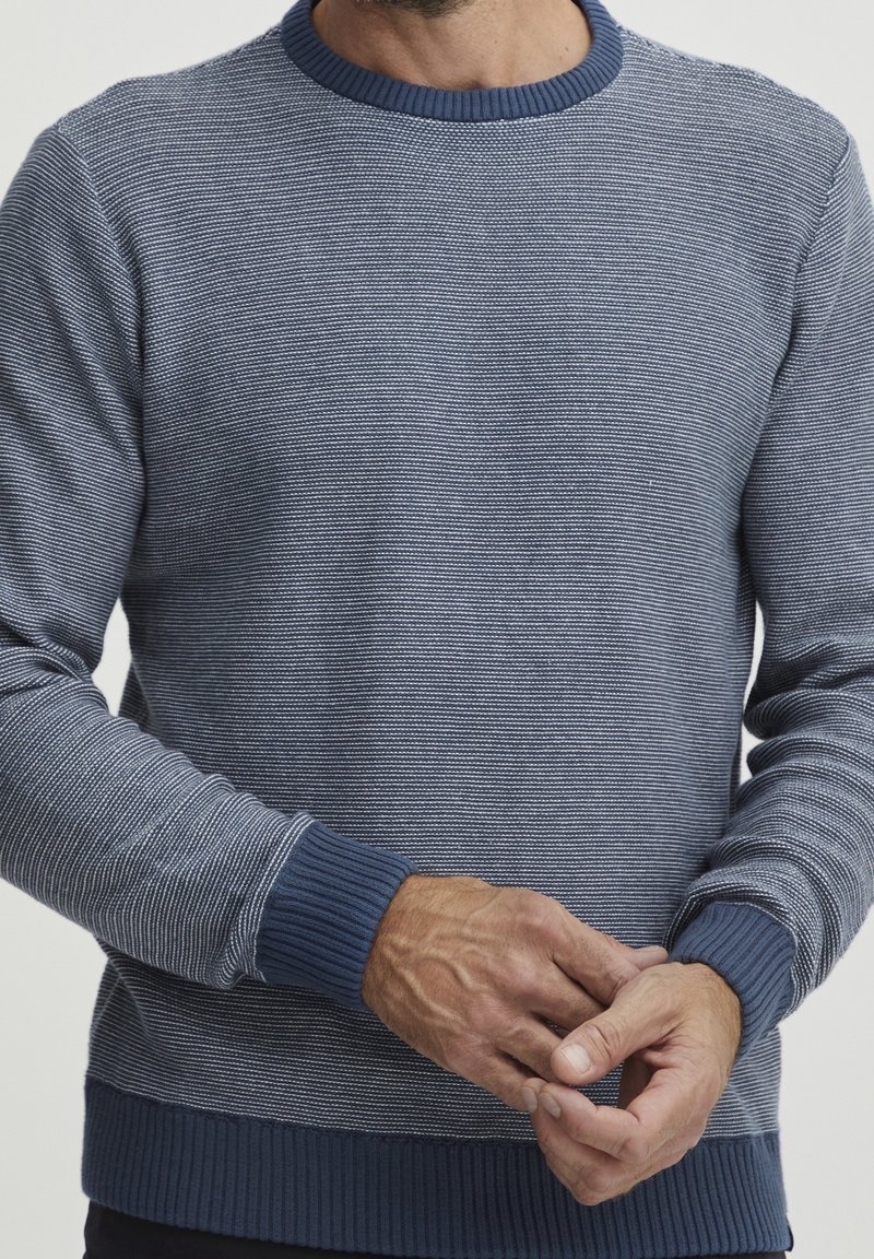 Homme portant un pull tricoté bleu et blanc texturé avec poignets et ourlet côtelés, les mains légèrement jointes à hauteur de taille.