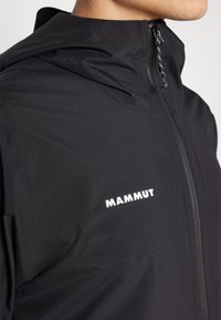 Crna jakna na zip od laganog materijala, s visokim ovratnikom i bijelim logotipom "MAMMUT" na lijevoj strani grudi.