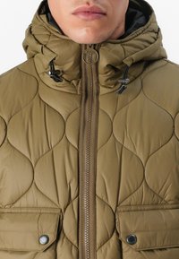 Jachetă puffer matlasată de un verde măslin, cu guler înalt, închidere cu fermoar și șnururi ajustabile. Prezintă buzunare frontale cu capse.