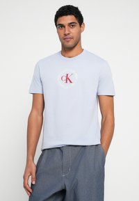 Jasnoniebieski t-shirt z krótkim rękawem i okrągłym dekoltem, z okrągłym czerwono-białym logo graficznym na piersi.