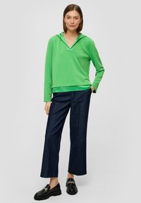 Zielona bluza z kapturem uszyta z miękkiego materiału, z dekoltem w kształcie litery V, zestawiona z ciemnoniebieskimi spodniami o szerokich nogawkach oraz czarnymi mokasynami z akcentami.